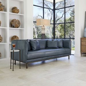 Featuring a blue leather sofa, a drink table with a wine glass on it. and behind an accent lamp and a ceiling to floor window