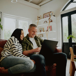 A couple on a cozy couch, he is looking at something in his laptop and she is next to him watching the screen, a beautiful plant next to the couch