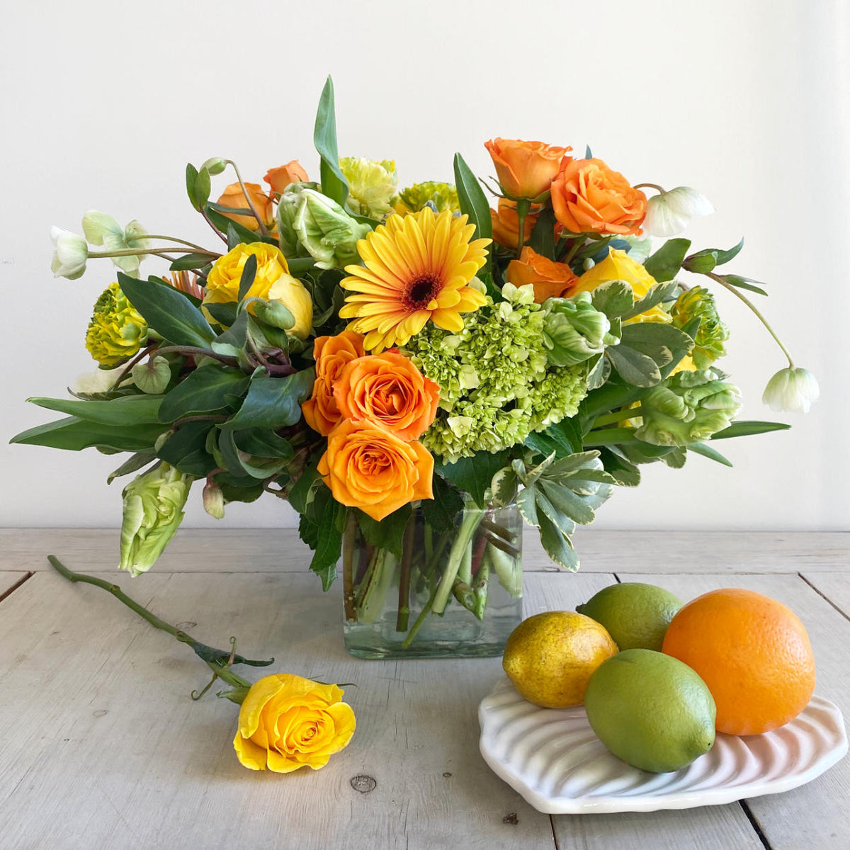 A beautiful citrus toned flower arrangement. A mix of lemon, limes, bright oranges, and stunning yellows. The arrangement features green Hellebore, green and white Parrot Tulips, orange crush Spray Roses, Kiwi Hydrangea, and large two-toned Gerbera Daisys. In a clear glass cylinder. next to it, a plate with lemons and limes, and there is a yellow rose over the table