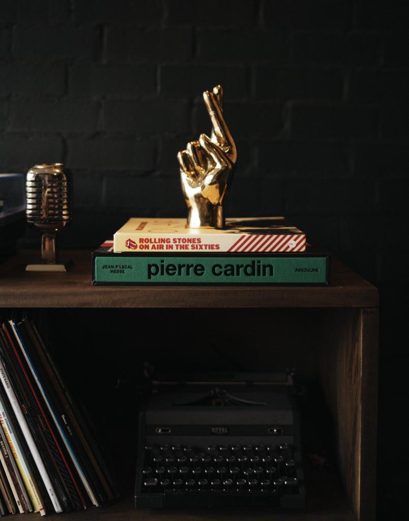 A sculpture of a life-size hand with crossed fingers in brass finish resting on a book that is on a console table next to a vintage typewriter and other decorative objects